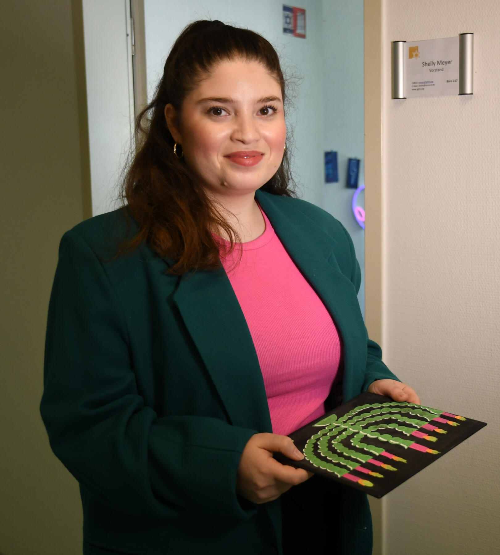 Shelly Meyer trägt einen grünen Blazer, ein pinkes Shirt und hält eine Zeichnung einer Chanukkia in der Hand. Sie steht vor ihrem ehemaligen Büro der jüdischen Gemeinde in Hamburg.